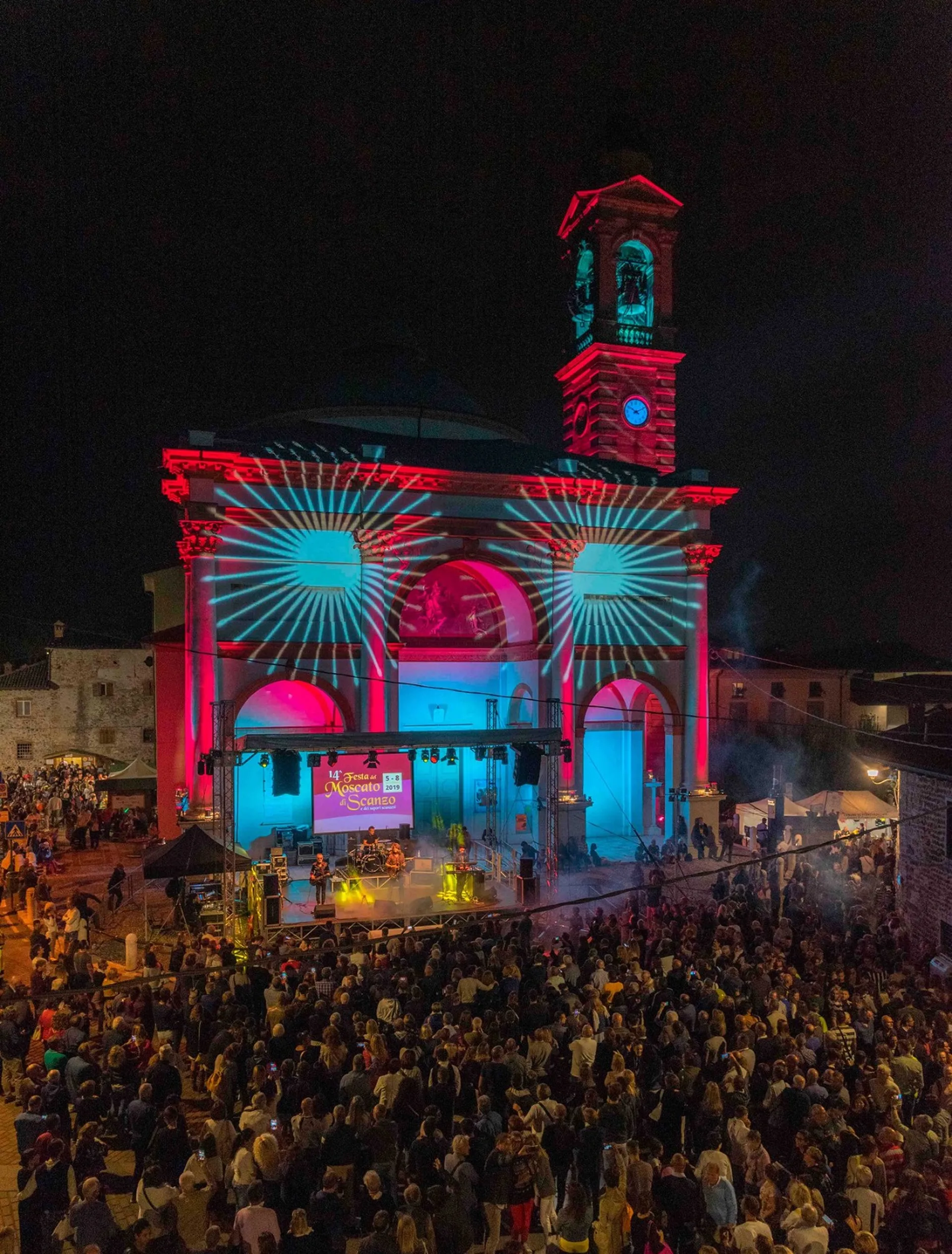 Chiesa di Rosciate illuminata con raggi ciano e magenta, folla della Festa del Moscato di Scanzo vista dall'alto — illuminazione architettonica SUONO E LUCE